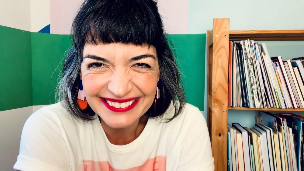 Flávia Scherner sorrindo, ao fundo estante de livros e parede branca com detalhes em verde e rosa.