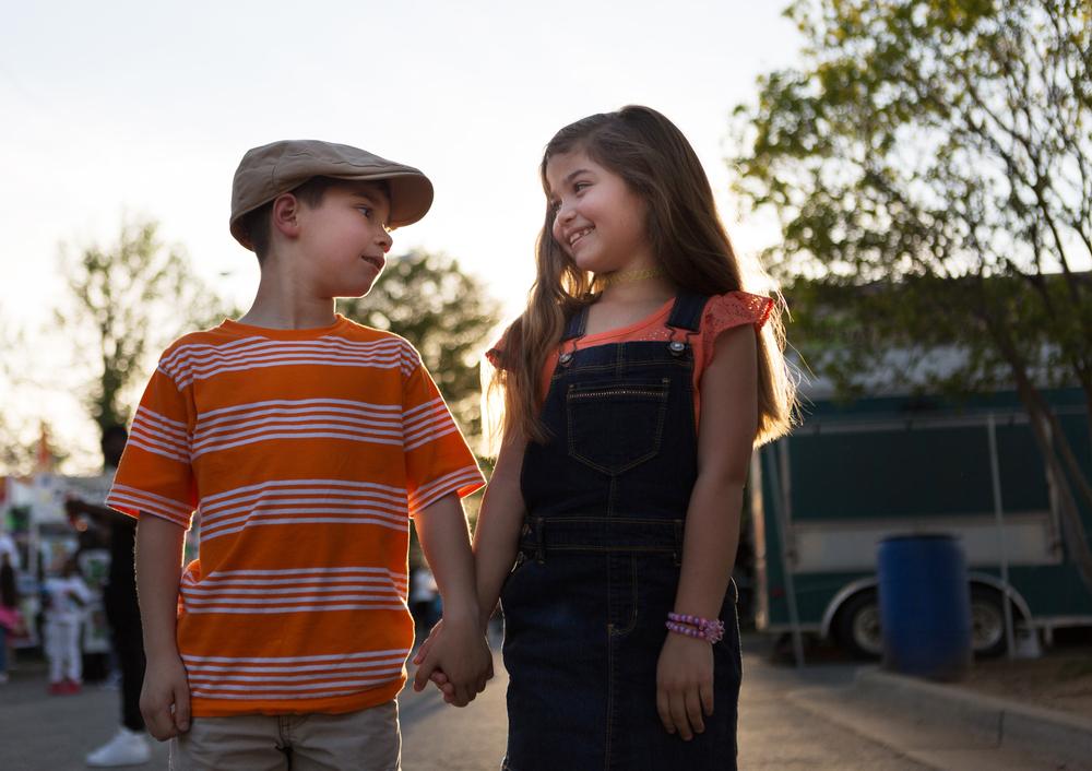  Imagem de um menino e uma menina de mãos dadas se olhando. O menino usa uma boina e uma camisa listrada laranja e branco e a menina está de cabelo solto, usando um macacão escuro e sorrindo