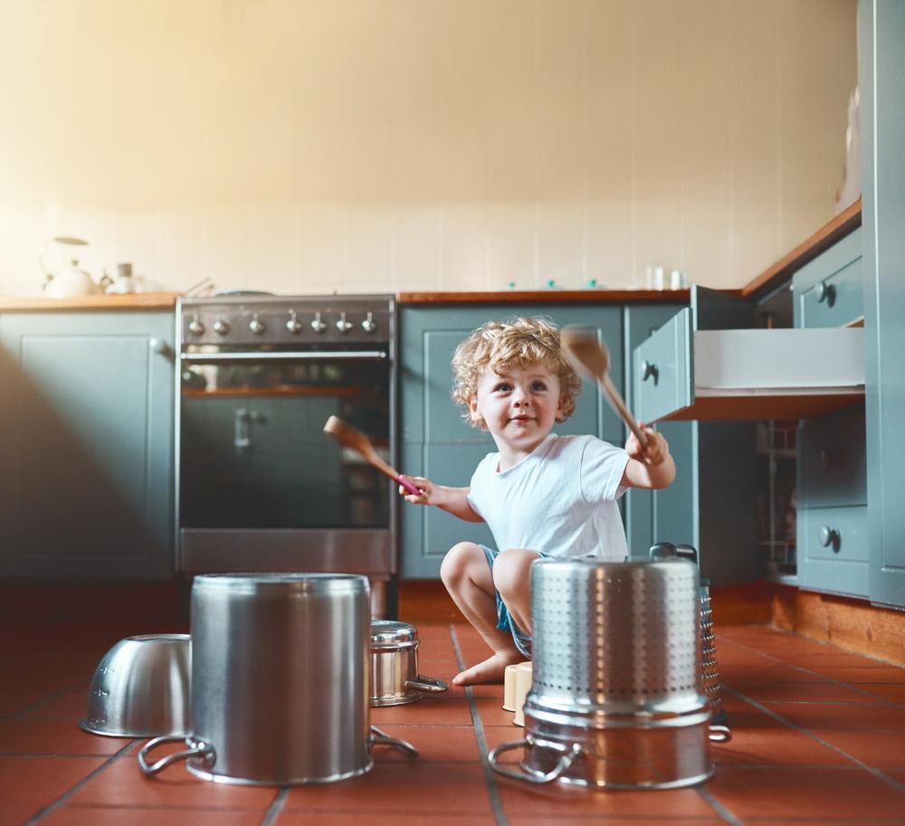 criança brincando com panelas e colheres de pau sentado no chão da cozinha.