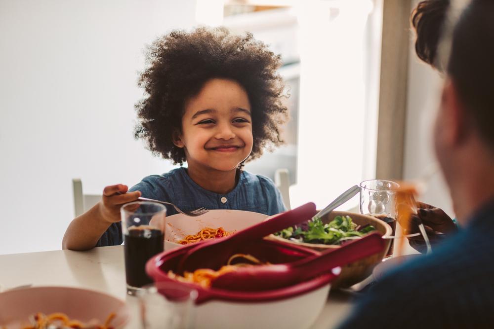 criança sentada a mesa no horário de almoço, sorrindo enquanto segura o talher na mão. Em sua frente estão os pratos com alimentos, incluindo uma salada verde, e há a presença de um adulto do outro lado da mesa.