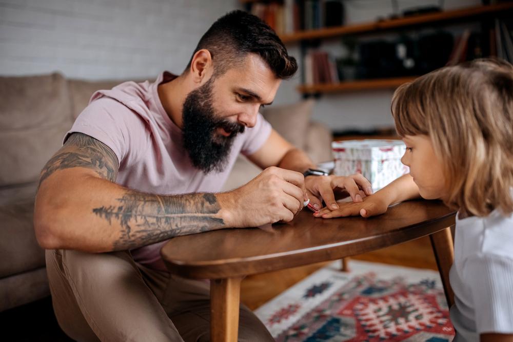 Imagem de um pai solo pintando com esmalte vermelho as unhas de sua criança. Ela está com a mão apoiada sobre uma mesa