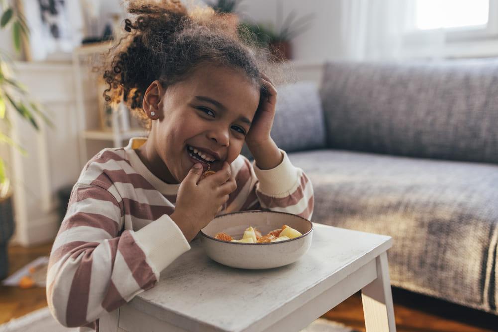 Foto de uma criança fazendo um lanche e comendo com as mãos uma vasilha de salada de frutas, um exemplo de alimento rico em vitamina c 