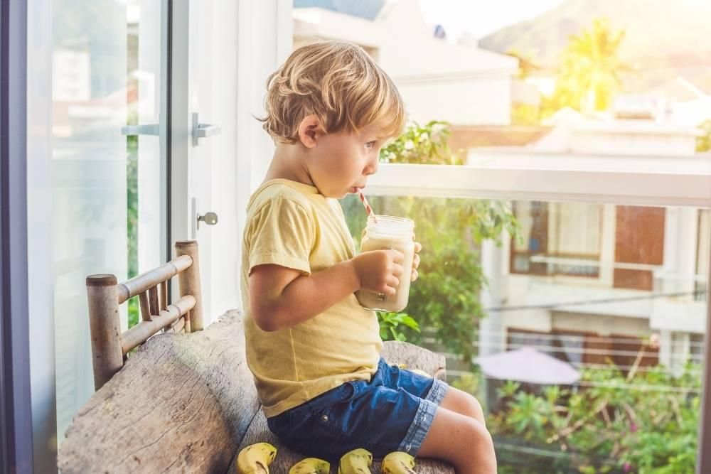 Criança sentada, segurando um copo com as mãos e tomando uma bebida feita com leite. Ao fundo, há uma janela de vidro com vista para outras casas. 
