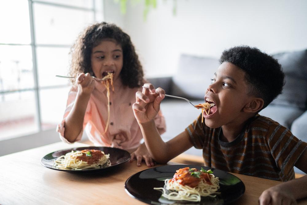 irmãos à mesa, comendo e se olhando na sala de casa