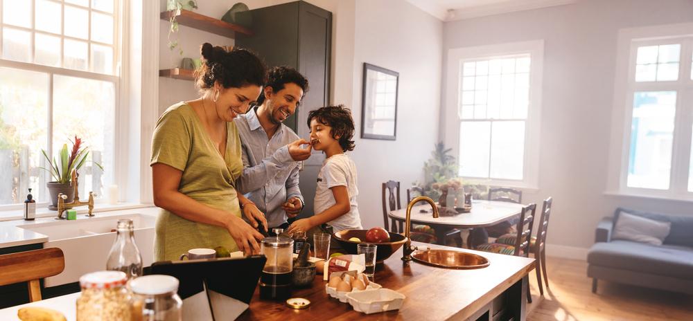 Uma família reunida na cozinha de sua sala. O pai oferece um cookie para o seu filho, enquanto a mãe ao lado acompanha a atividade familiar. A foto remete ao tema cookie caseiro