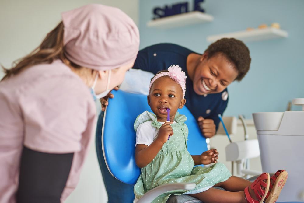 Uma menina está sentada na cadeira de uma dentista, com a escova de dentes na boquinha, observada pela sua mãe e a profissional. O cenário nos remete a importância da saúde bucal infantil