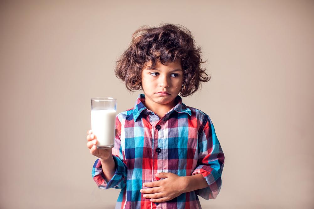 Imagem de uma criança desconfortável, com um copo de leite em uma mão e outra sobre a barriga em uma postura que remete aos efeitos da intolerância à lactose.