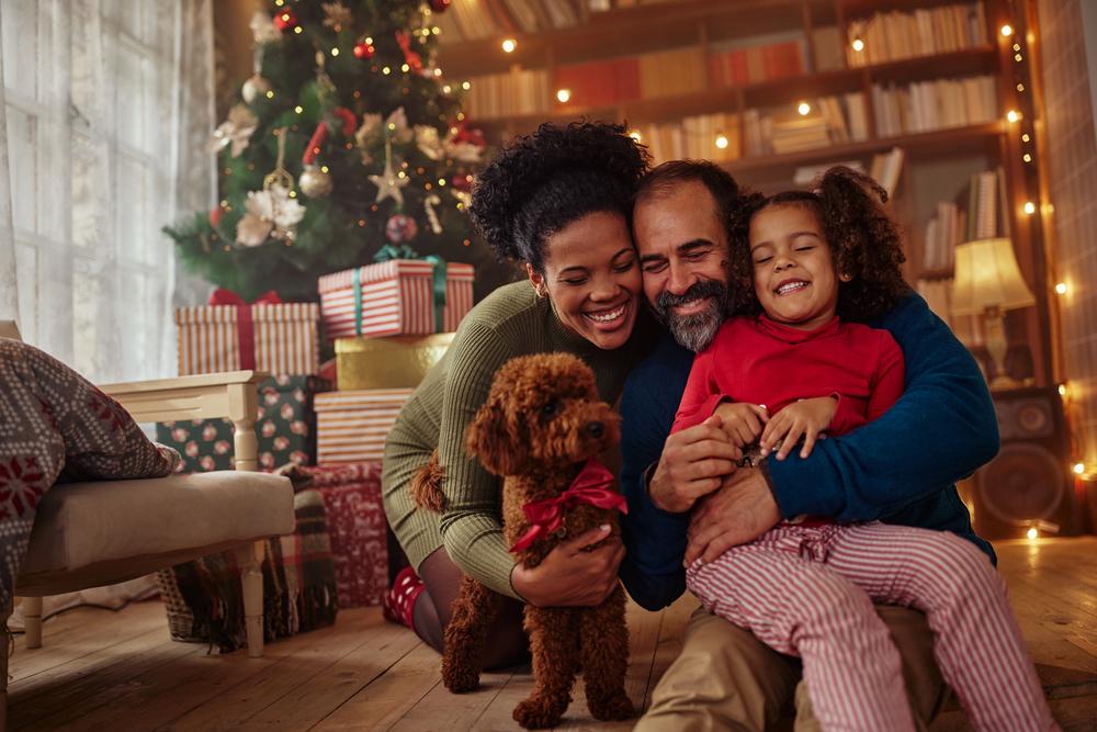 Homem e mulher abraçados com a filha e um cachorrinho no chão, na sala de casa em frente a uma árvore de natal