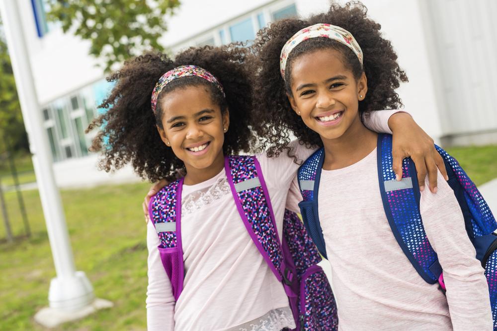 Duas irmãs gêmeas se abraçando lado a lado, sorridentes, com mochila nas costas, ao ar livre em frente a escola.