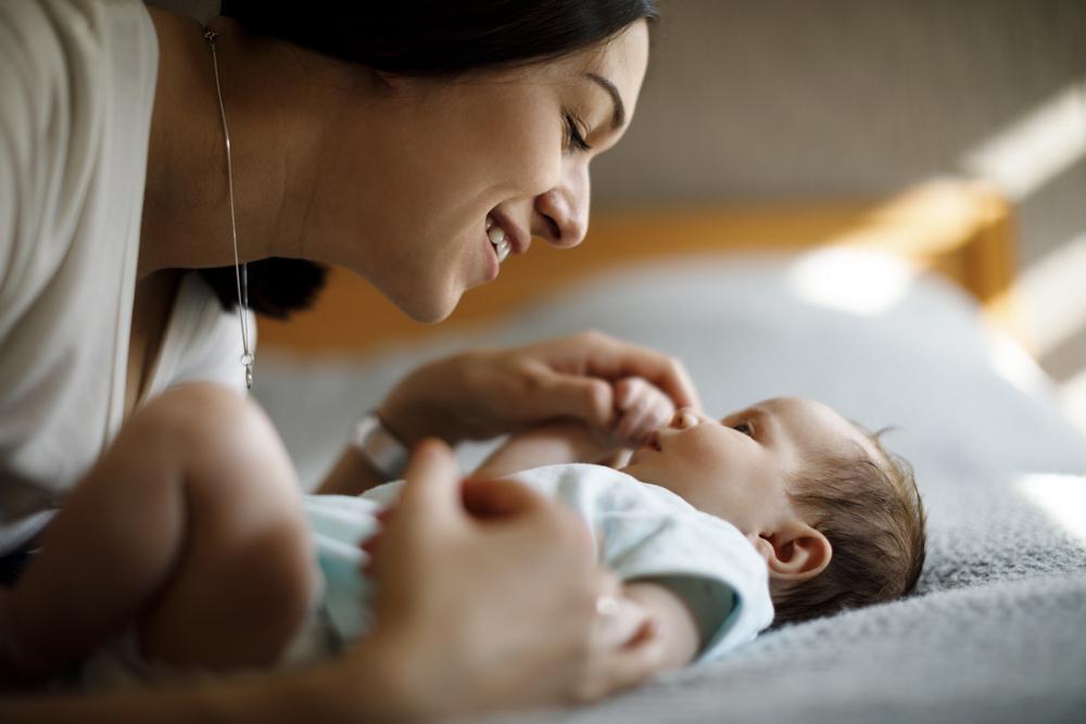 Imagem de uma mãe acariciando um bebê deitado. O carinho e a troca de olhares entre eles ajuda a fortalecer o vínculo entre os dois