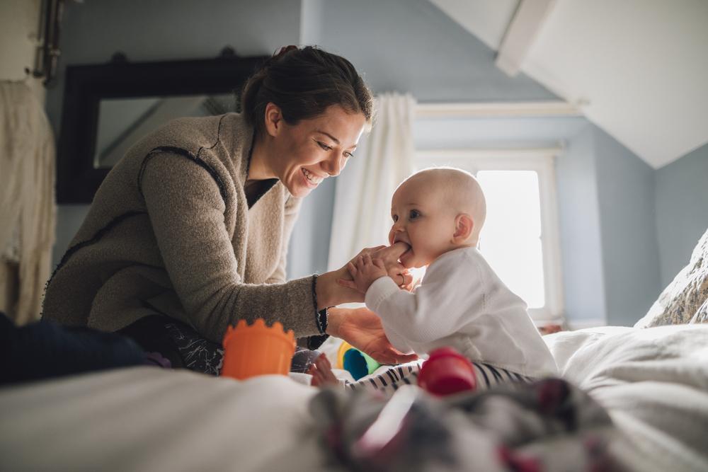 Um bebê está sentado na cama, mordendo o dedo da sua mãe que está na sua boca. A imagem remete ao tema de cárie formada pelo uso da mamadeira