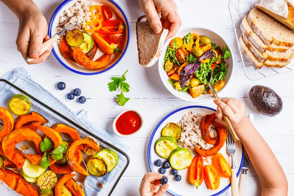 Foto tirada de cima de uma mesa com a alimentação familiar. Há mãos de adulto e de criança e elas se servem de diversos alimentos alis dispostos: pães, frutas, cereais e legumes.