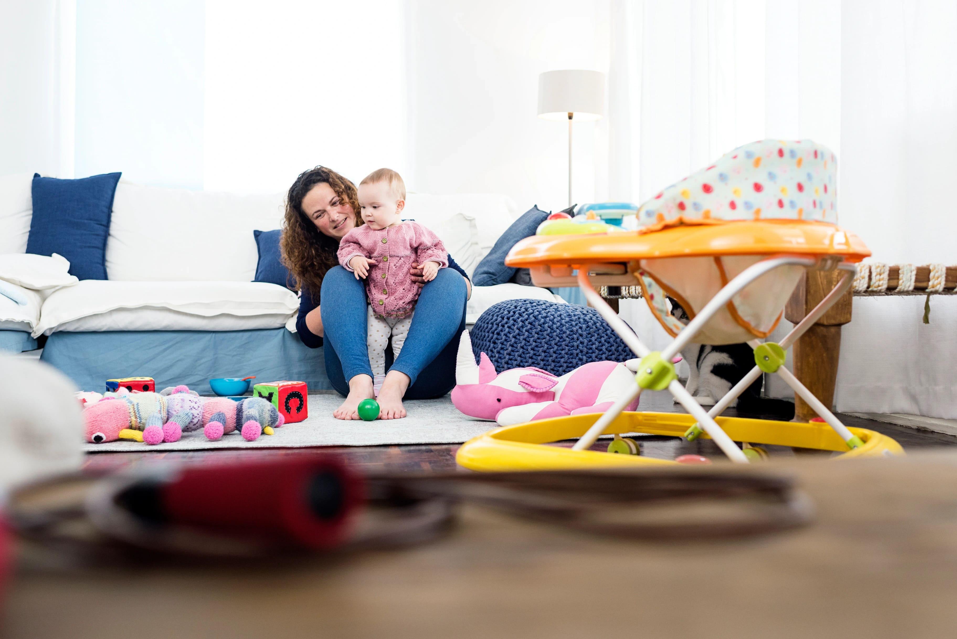 Ao fundo temos uma mãe brincando com um bebê e o estimulando a ficar em pé, ao lado temos brinquedos e um andador infantil