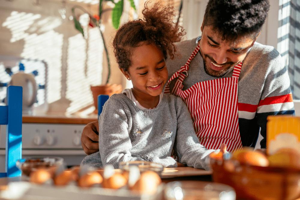 A relação de pai e filho é ilustrada por uma imagem onde a menina é abraçada pelo pai, que veste avental, enquanto eles olham para uma mesa de café da manhã com alguns alimentos.