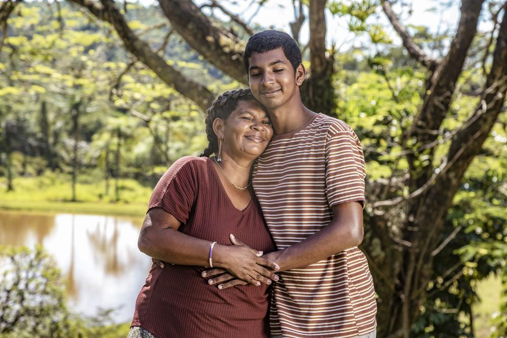 Mulher negra de cabelo preto vestindo uma roupa vermelha abraçando seu filho negro que veste uma roupa listrada com detalhes marrom claro e branco, no fundo um lago e árvores em volta. 