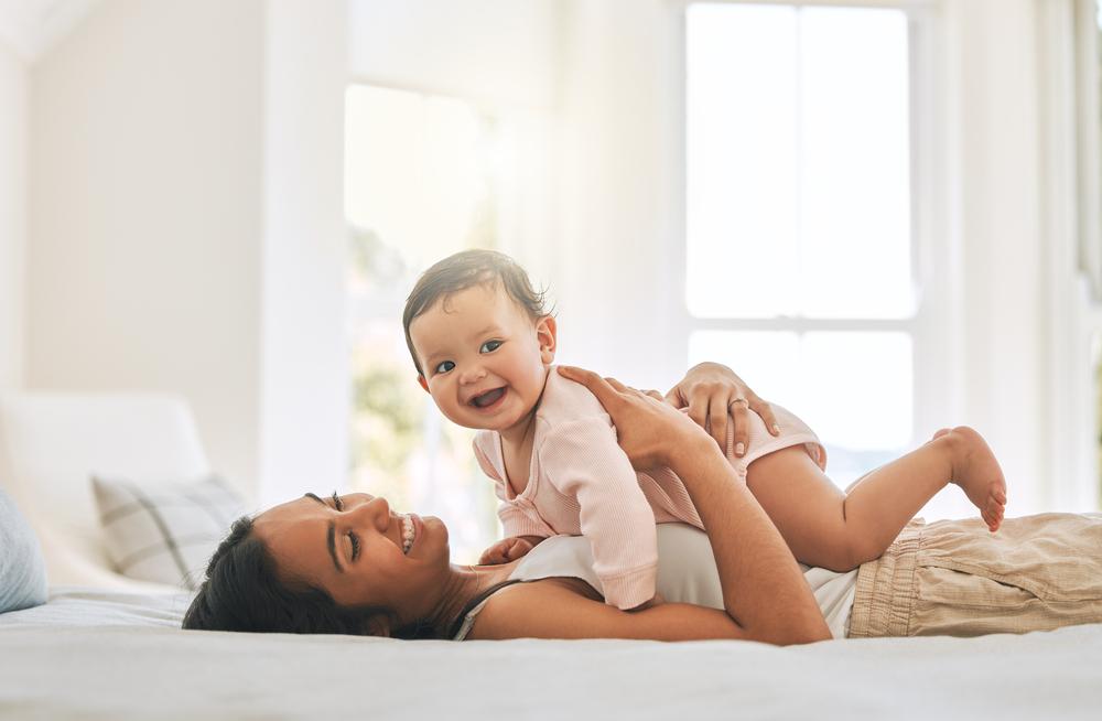 Mãe morena de cabelo preto liso deitada na cama de barriga para cima segurando seu bebê 