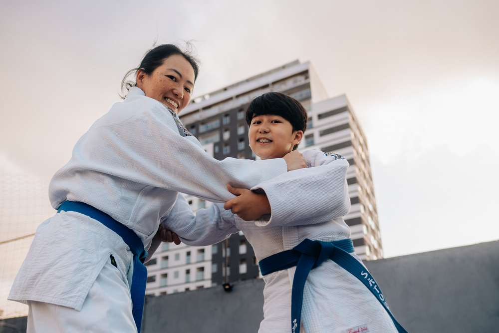 Mãe e filho vestindo kimono com uma faixa azul fingindo estarem lutando enquanto olham para a câmera 