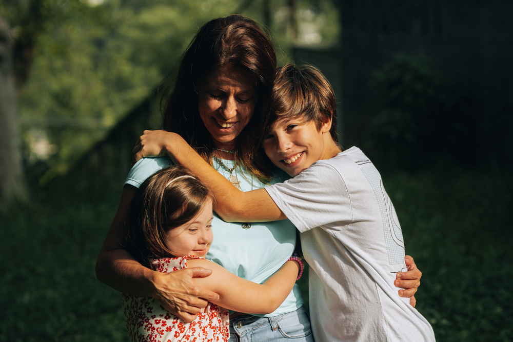 Uma mãe branca vestindo uma blusa azul esverdeado abraçando seus filhos, do lado direito um menino branco e loiro vestindo uma blusa branca e do lado esquerdo uma menina com síndrome de down utilizando um vestido branco com detalhes vermelhos