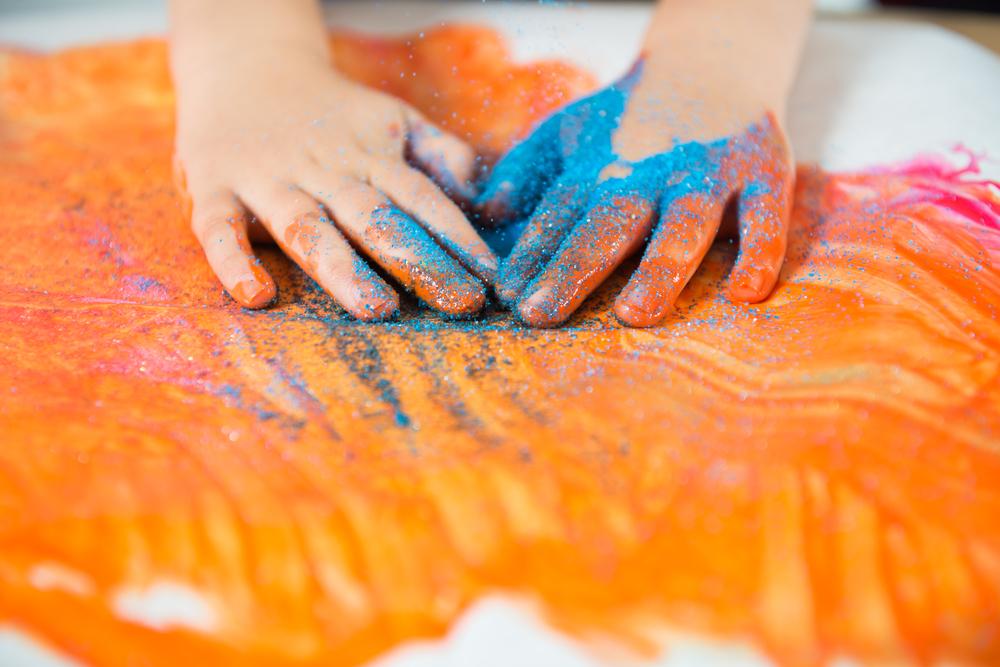 Imagem das mãos de uma criança, sentindo a textura de um glitter e de tintas. Ao fundo, um cartaz branco com algumas pinturas na cor laranja, rosa e glitter azul.