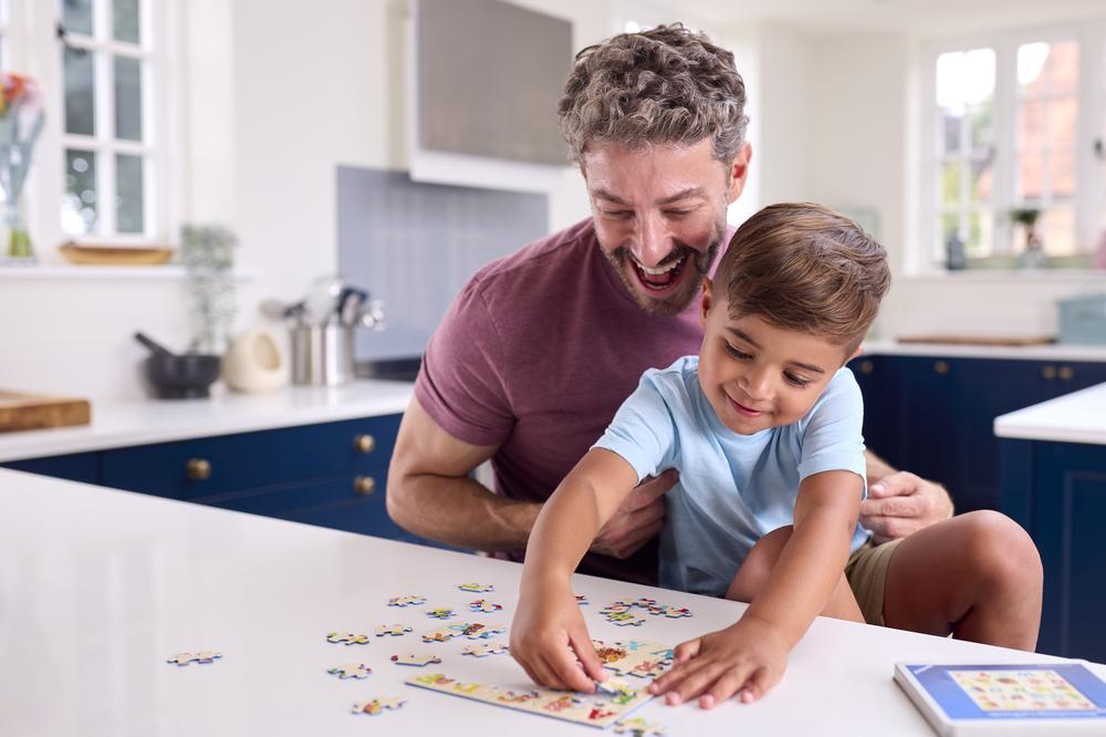  Imagem de pai e filho montando um quebra-cabeça. Ao fundo, uma cozinha com armários na cor azul e bancada branca.