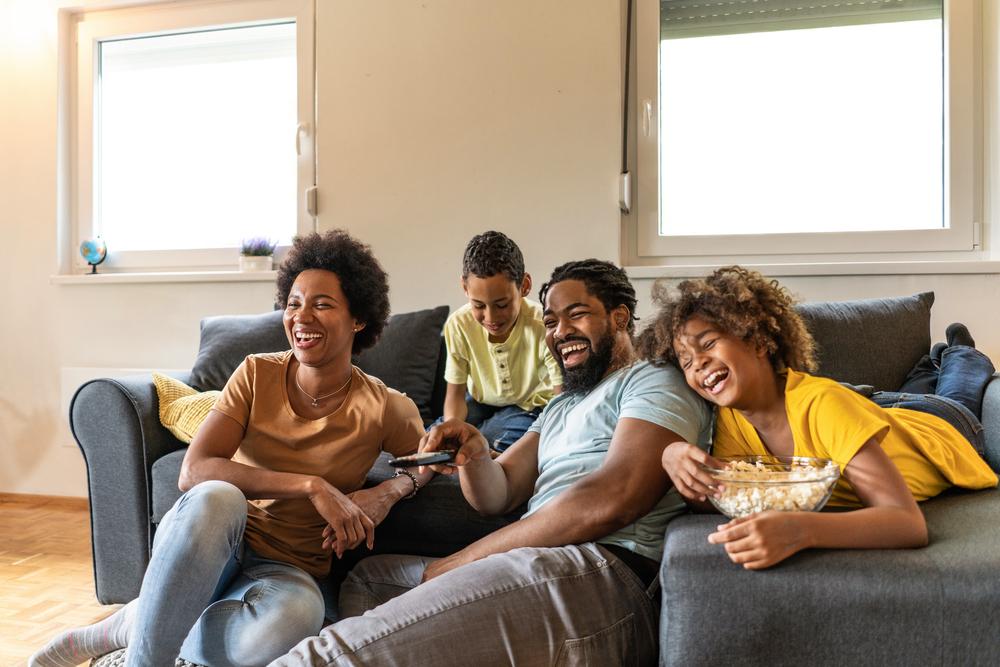 Imagem de uma família assistindo a filmes infantis. Mãe e pai estão sentados no chão e os filhos no sofá. Todos estão gargalhando.