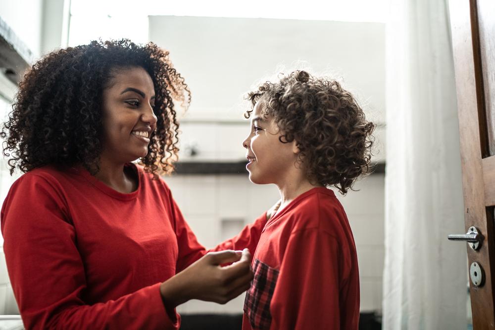 Mulher sorrindo com seu filho, ambos usando um pijama vermelho e conversando. Ao fundo, um quarto.