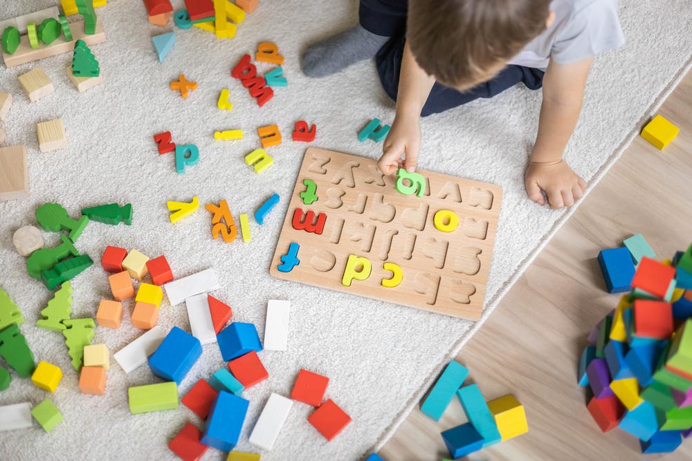 Em um piso de madeira com um tapete cinza, está sentado um menino,vestindo uma camiseta branca. Ele está brincando com um tabuleiro de encaixar letras, enquanto segurando uma delas. Há letras e outras peças coloridas espalhadas ao seu redor.