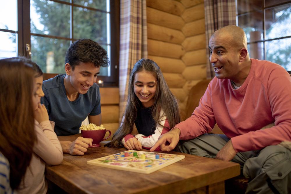 Dentro de uma casa, todos sentados em volta de uma mesa de madeira, está uma família composta por um homem, uma mulher, duas meninas e um menino. Com sorrisos e brincadeiras, eles estão jogando um jogo de tabuleiro.
