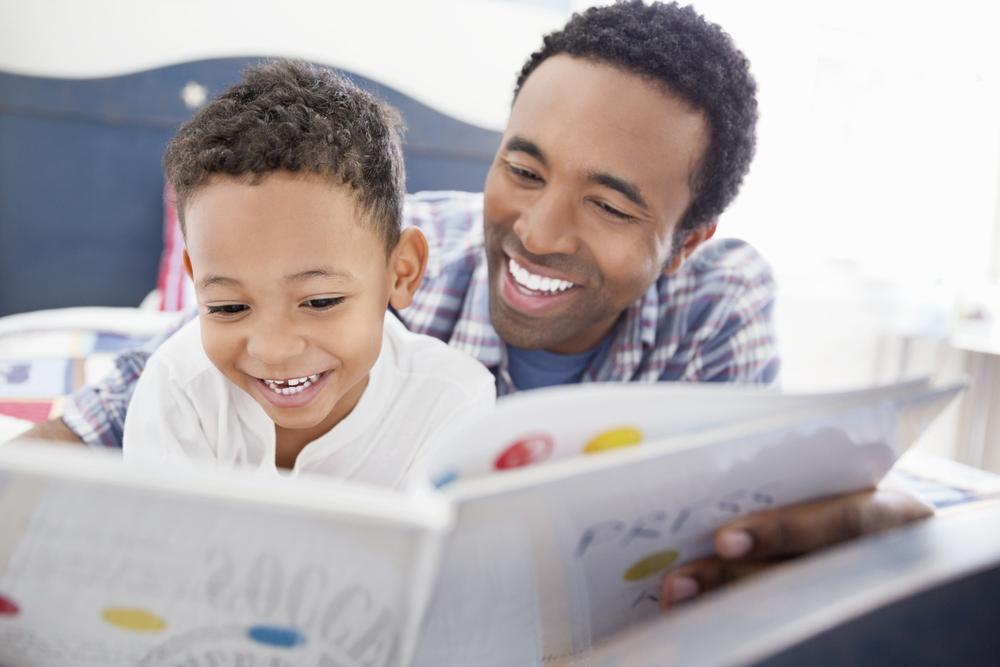 Deitados em uma cama, um homem, vestido com uma camisa quadriculada azul, está lendo um livro com um menino, que veste uma camisa branca. Com o livro aberto, o menino e o homem estão sorrindo. 