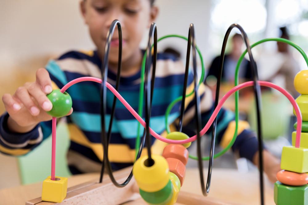 Em uma sala, com pessoas ao fundo, um menino, que está vestindo uma camisa com listras azuis, brancas, amarelas e marrons, está sentado à mesa, brincando com um brinquedo colorido, com formas geométricas.