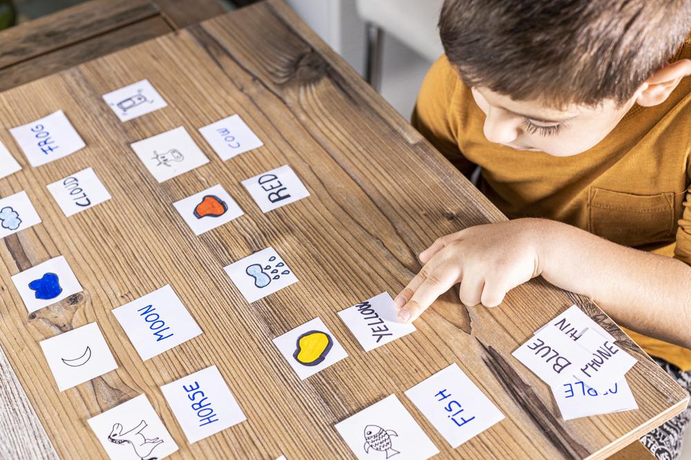 Sentado à mesa, está um menino vestindo uma blusa amarela. Sobre a mesa, há várias cartas com imagens de cores e animais, juntamente com seus respectivos nomes. O menino está associando os nomes às imagens correspondentes.