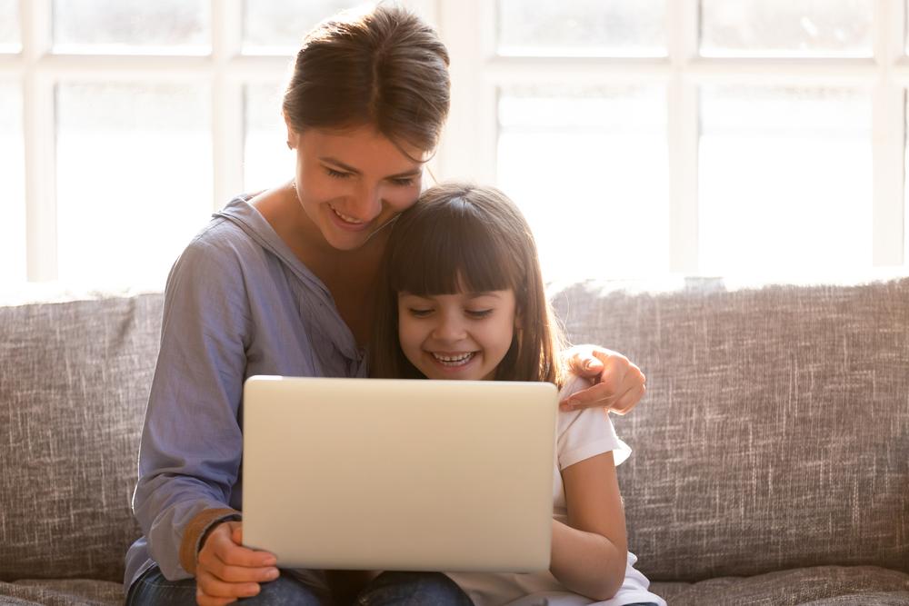 Sentadas em um sofá cinza, estão uma mulher e uma menina. A mulher está segurando um tablet. Sorridentes, ela e a menina assistem a um vídeo no dispositivo.