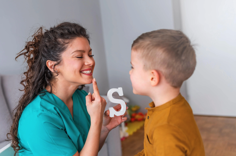Uma mulher sorrindo, apontando para a própria boca e segurando um objeto em forma de letra “S”, enquanto uma criança observa e tenta imitar a ação da mulher.