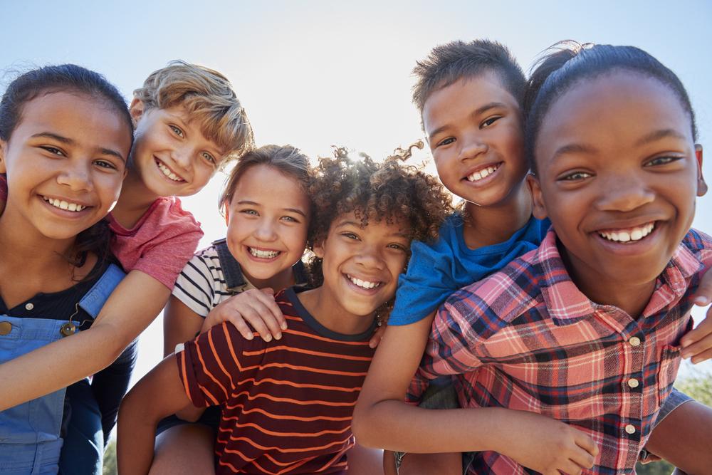 Ao ar livre, em um dia ensolarado, seis crianças - três meninas e três meninos - estão abraçadas entre si, sorrindo enquanto brincam.