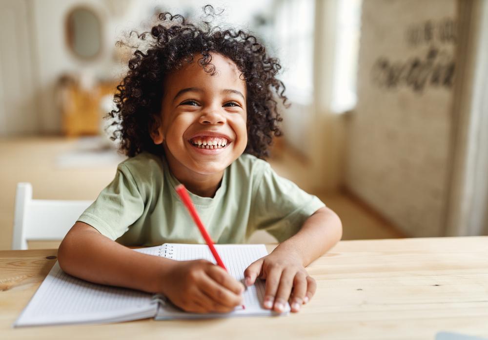 Descrição: criança sorrindo, segurando um lápis e um caderno para escrever. ao fundo, uma sala como ambiente.