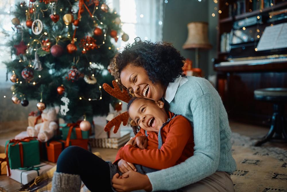 Em uma sala enfeitada para o Natal, está uma mulher e uma criança. Elas estão sentadas no chão, abraçadas e rindo.